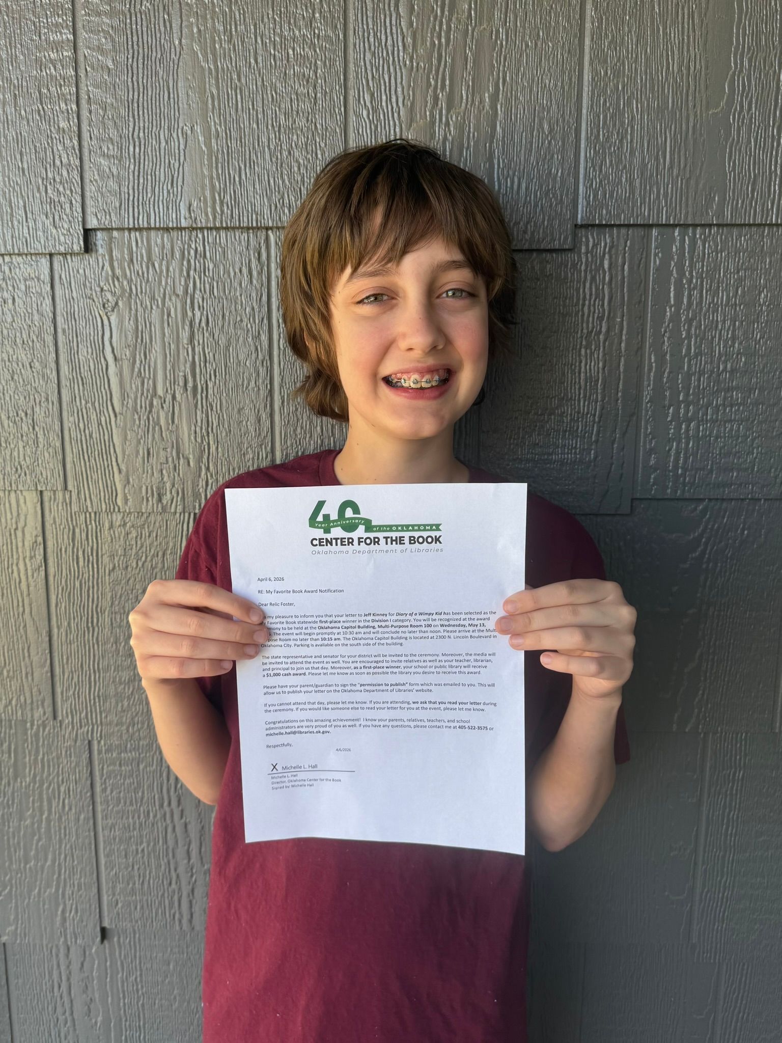 Smiling person in maroon shirt holding a certificate in front of a gray wall