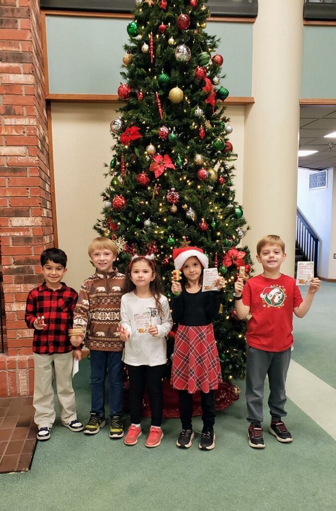 Five children stand in front of a decorated Christmas tree, holding gifts and smiling, in a brightly lit room.