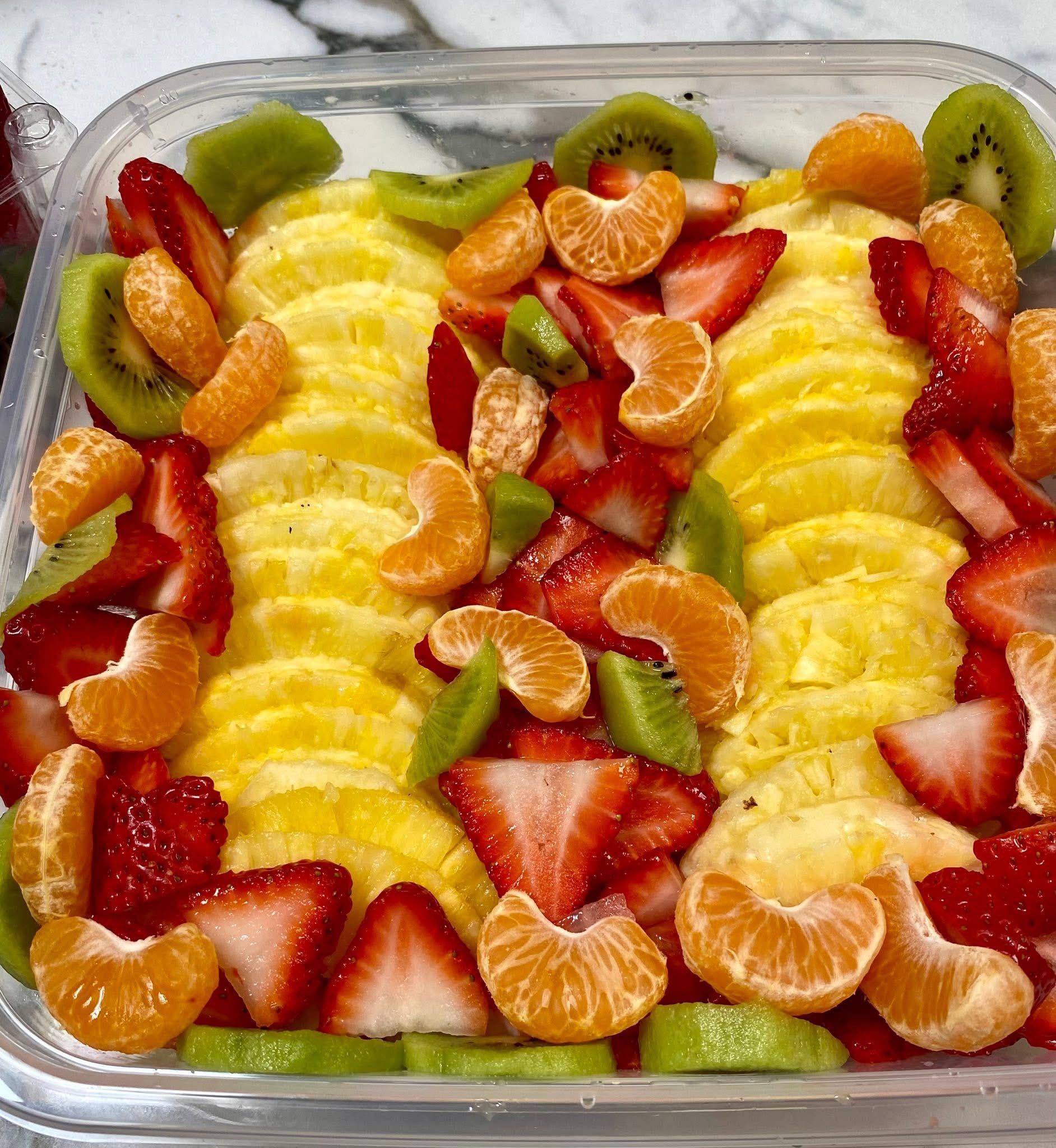 A plastic container filled with pineapple , strawberries , oranges , kiwi and mandarins.