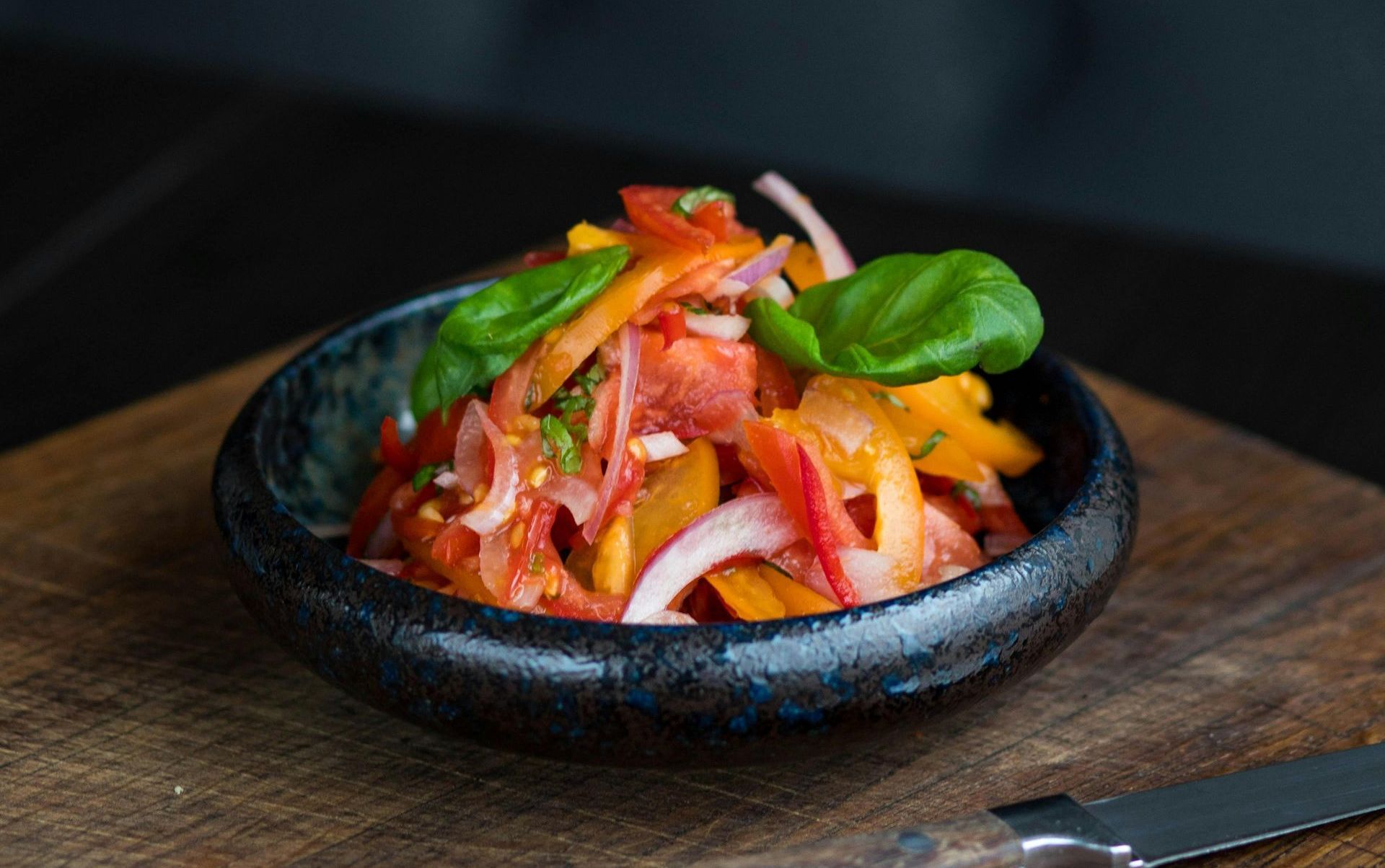 A bowl of salad with tomatoes , peppers , onions and basil on a wooden cutting board.