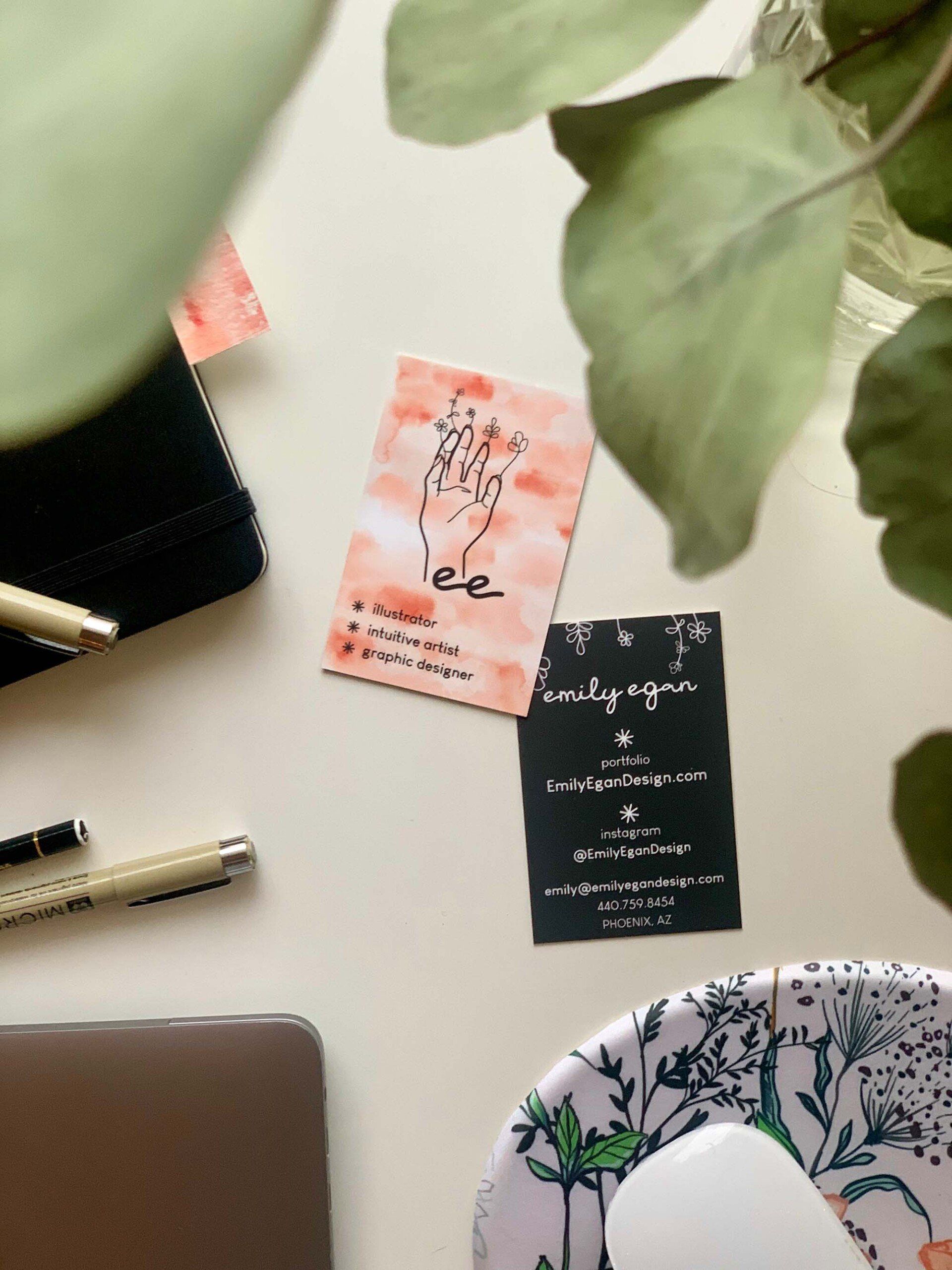 photo of Emily Egan's business card front and back with a notebook, Mircon pens, a pencil, the top right corner of a macbook, and part of a mouse and mousepad all showing on a white desk taken from an angle showing a eucalyptus plant in the foreground.