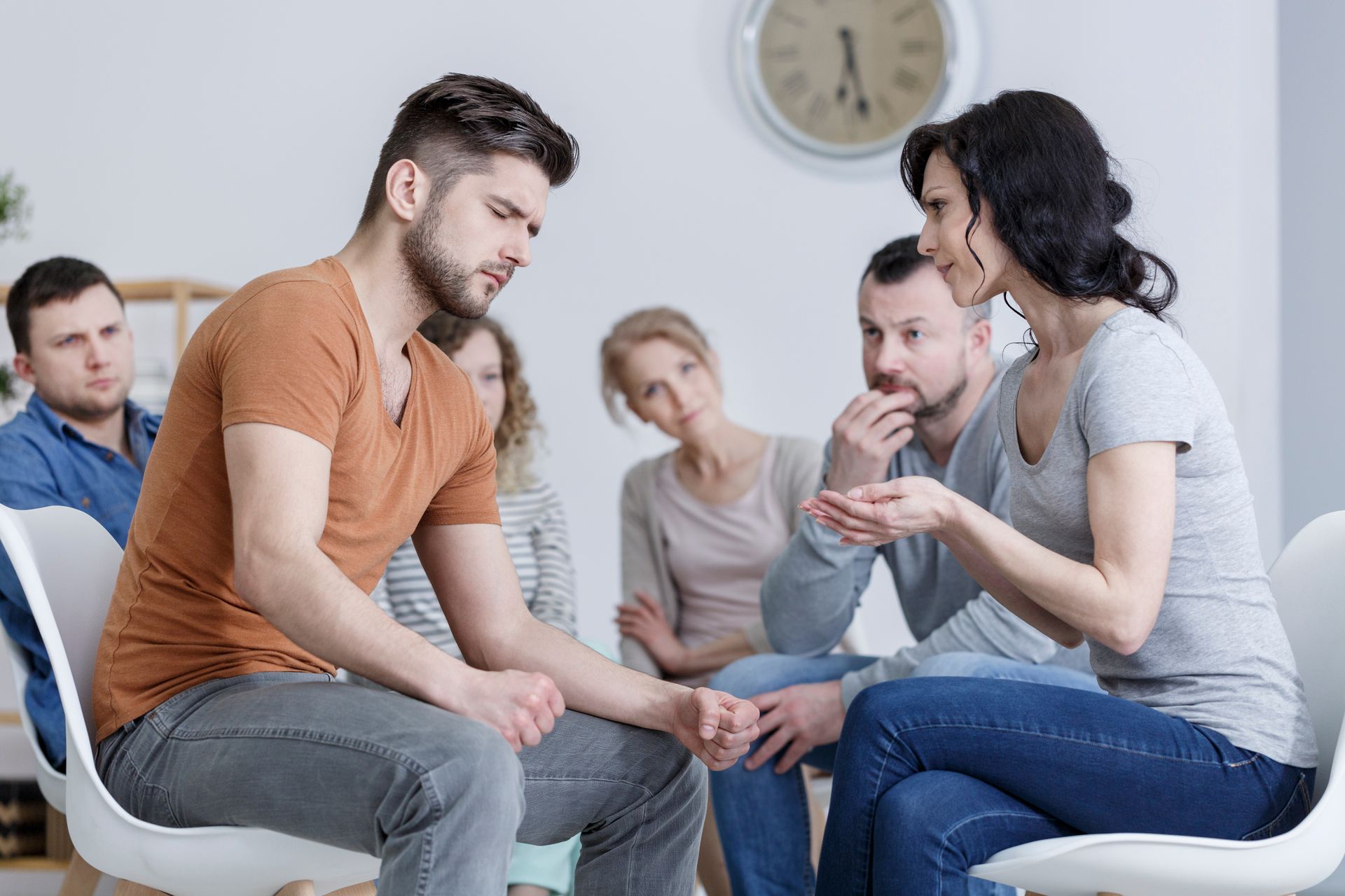 A group of people sits in a circle for a support group, with two individuals in the foreground talking to each other.