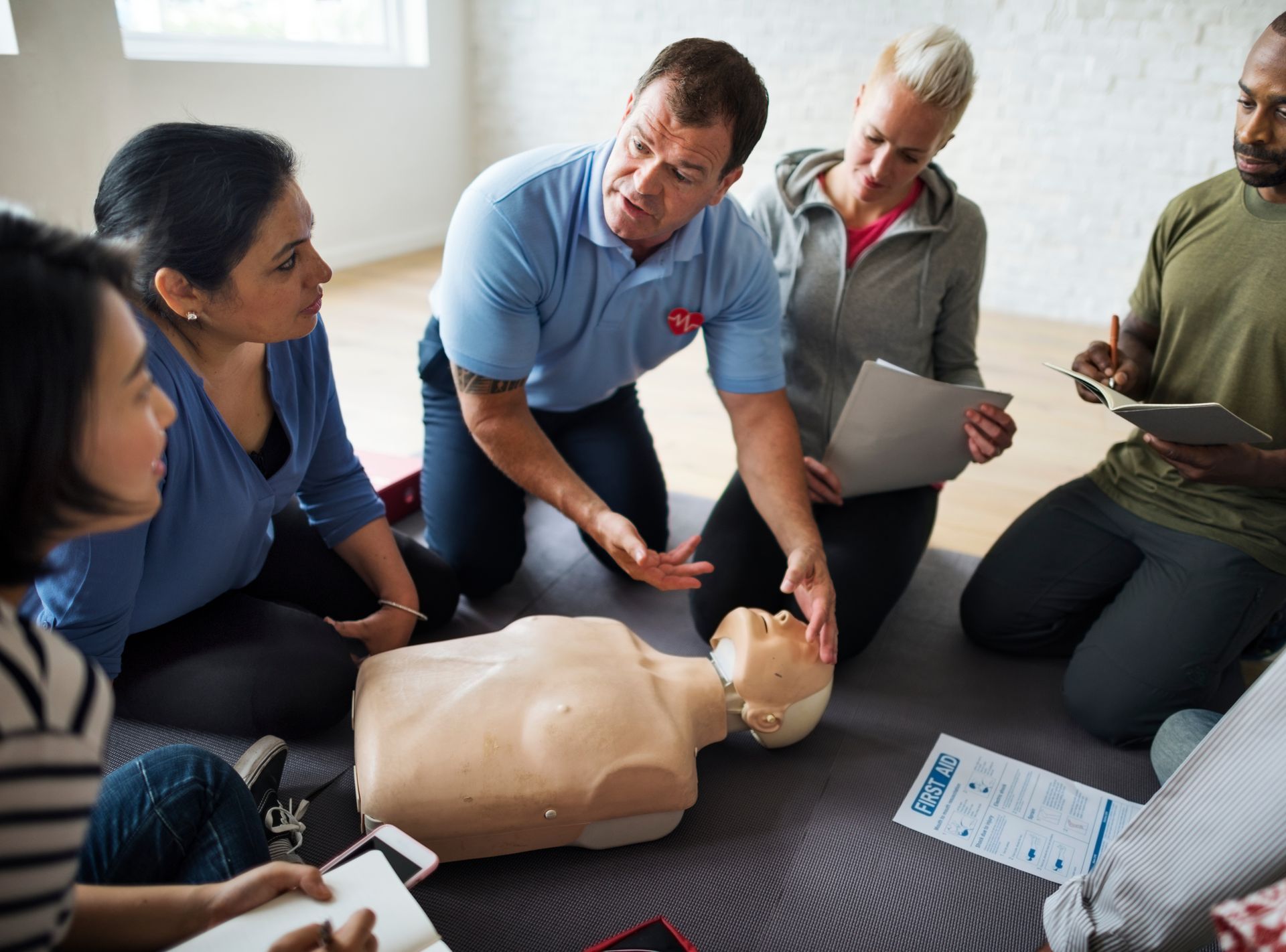 An instructor demonstrates CPR techniques on a mannequin to a group of participants sitting in a circle.