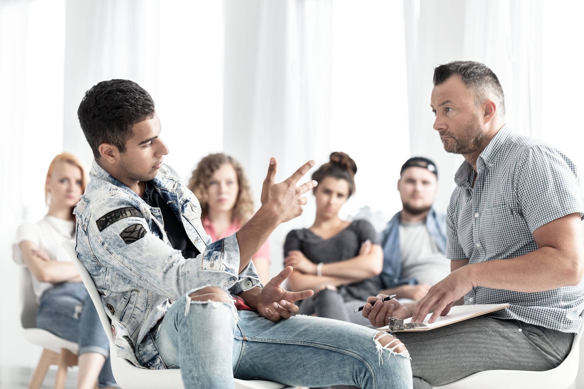A person gestures while speaking to a group leader holding a clipboard, with others sitting and listening in the background.