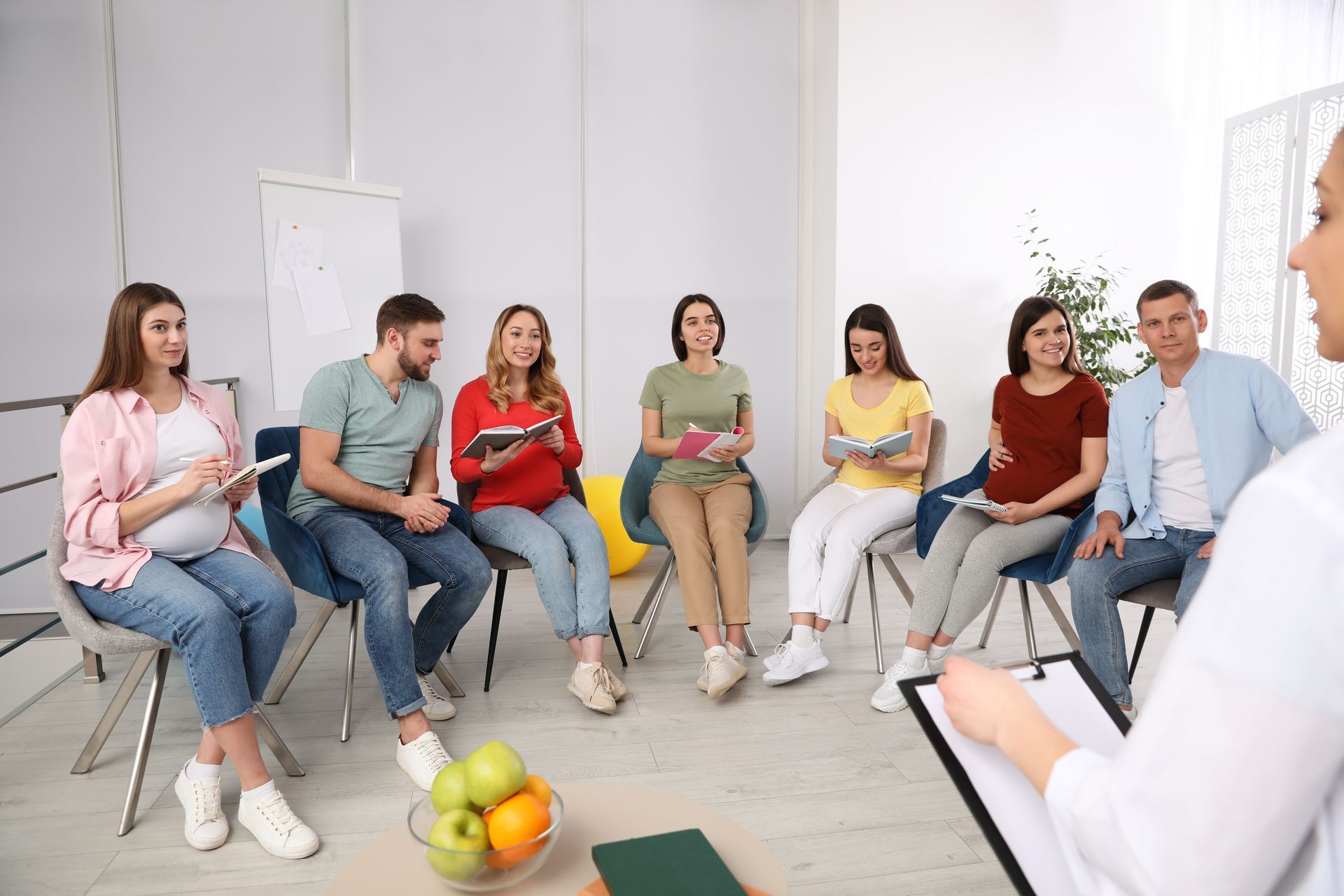 A group of people sits in a circle for a meeting or class while a facilitator in the foreground holds a clipboard.