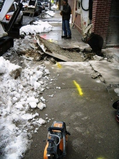A man is standing on a sidewalk looking at a hole in the ground
