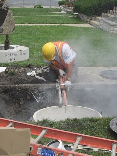 A construction worker is cutting a hole in a manhole cover with a chainsaw.