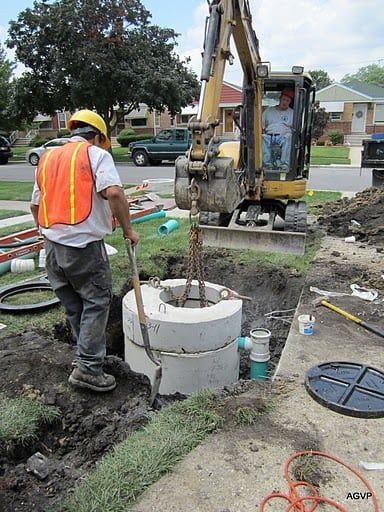 A man in an orange vest is standing next to a man in a yellow excavator.