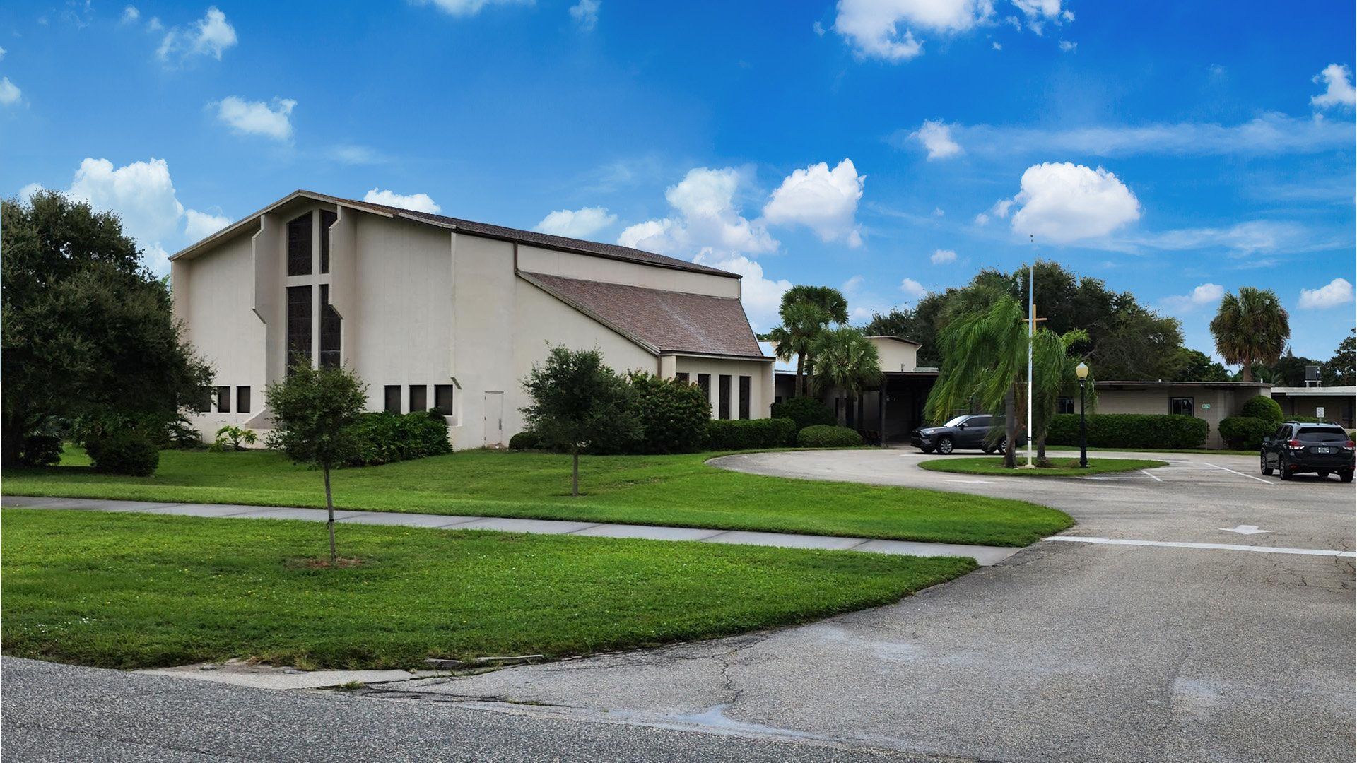 A large white building with a cross on the side is surrounded by green grass and trees.