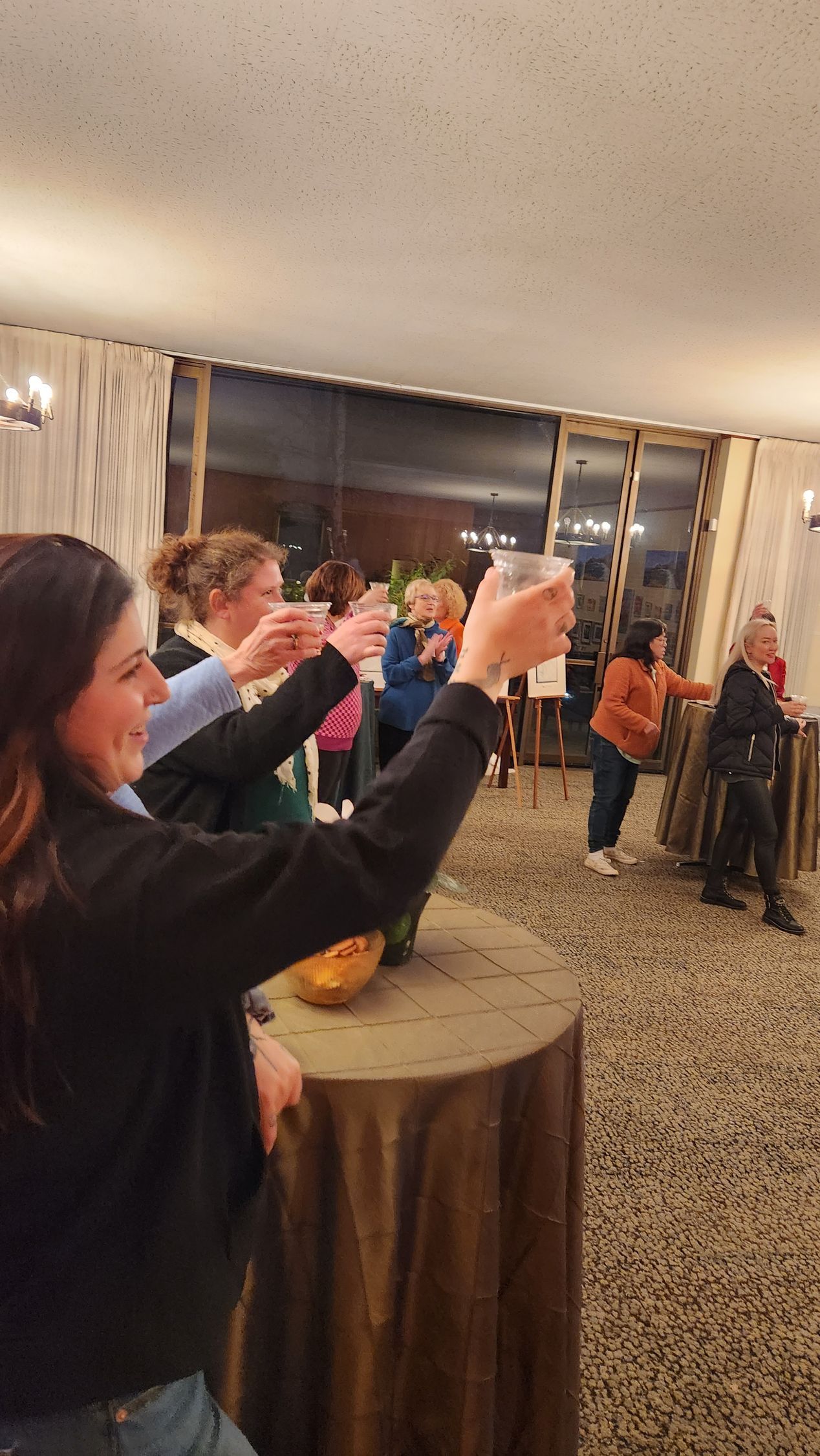 Many adults in the lounge at high-top tables raising their glasses in a toast