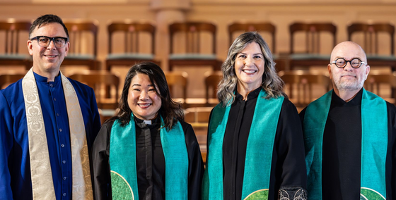 A picture of Calvary's three pastors Victor, Joann, and Marci, and Music Director, Michael in front of the stained glass windows on the 2nd/balcony level of Calvary