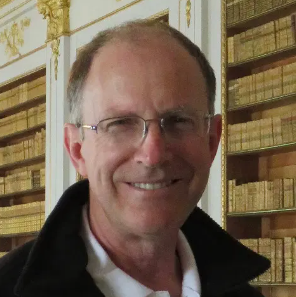 A picture of David Foster. He wears thin, wire-framed glasses. He is standing in front of an elegant book case that is built into a wall. It is white with gold trim