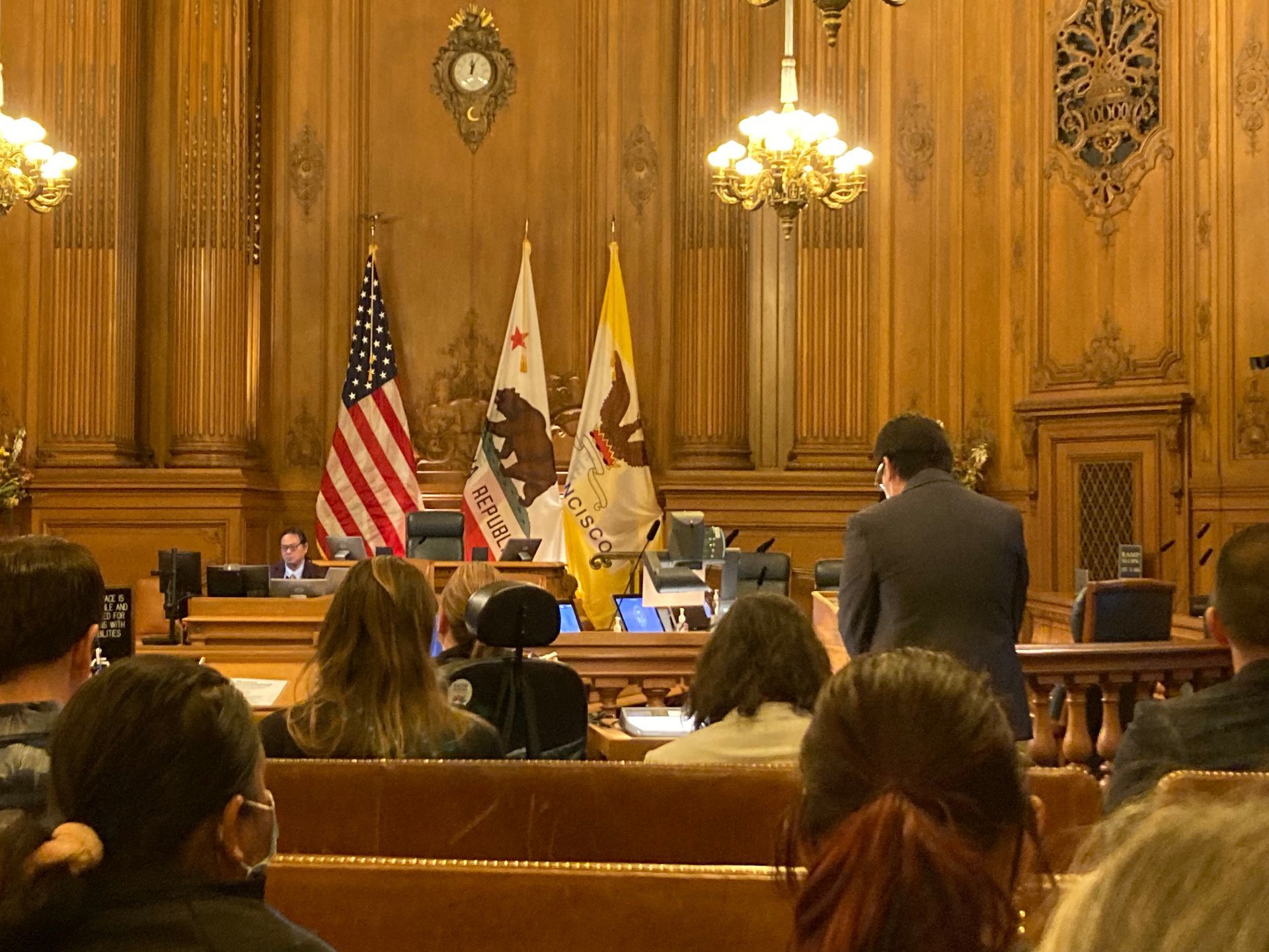 A shot of the courtroom, it has tan walls with lots of ornamental designs. There are 3 flags on the wall, the US flag, California Republic, and San Francisco's flag. 