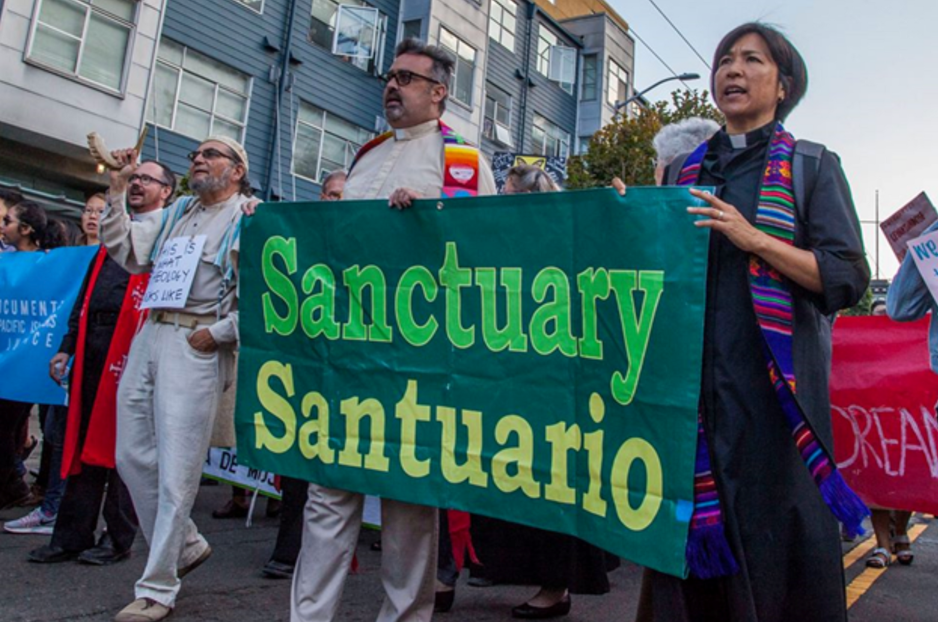 A crowd of marchers holding a banner that says Sanctuary/Sanctuario en español