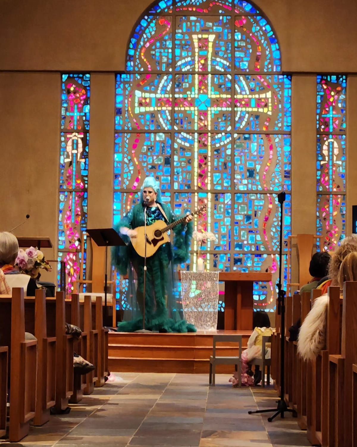 Flamy Grant, drag queen and drag artist, on stage in the Chapel with a guitar and behind a mic stand. She is wearing a glorious dark teal gown with lighter teal hair. There are many people in the seats.