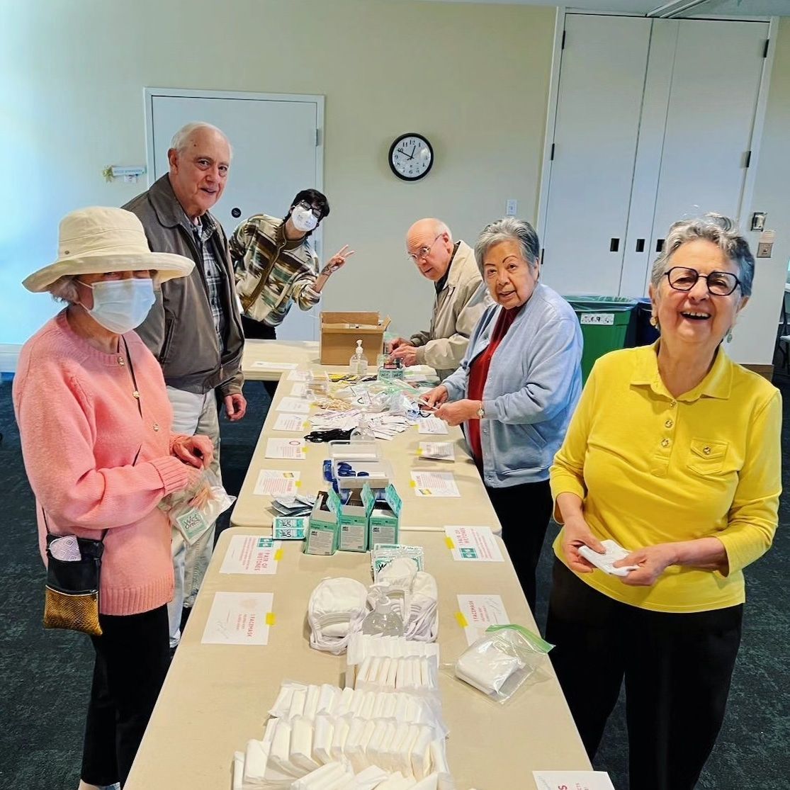 A group of Senior Adults packing hygiene kits for the Hope Center. There are various hygiene items on the table. Someone is giving a peace sign in the back.