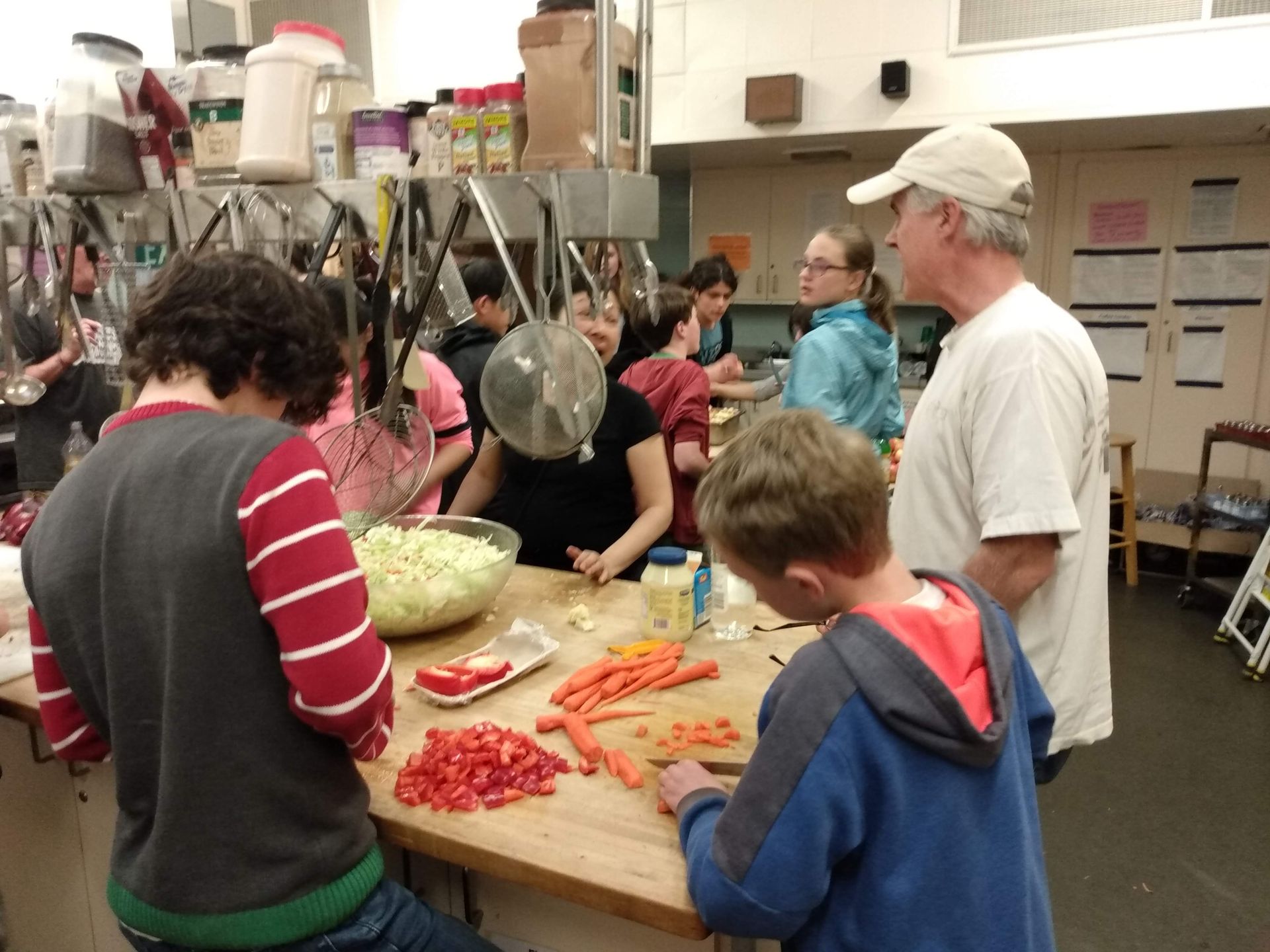 A group of Calvary folks chopping vegetables at a large commercial kitchen for one of our volunteer service events. Kitchen utensils hang from a rack on top of a large wooden island where everyone is cutting carrots, tomatoes, etc.