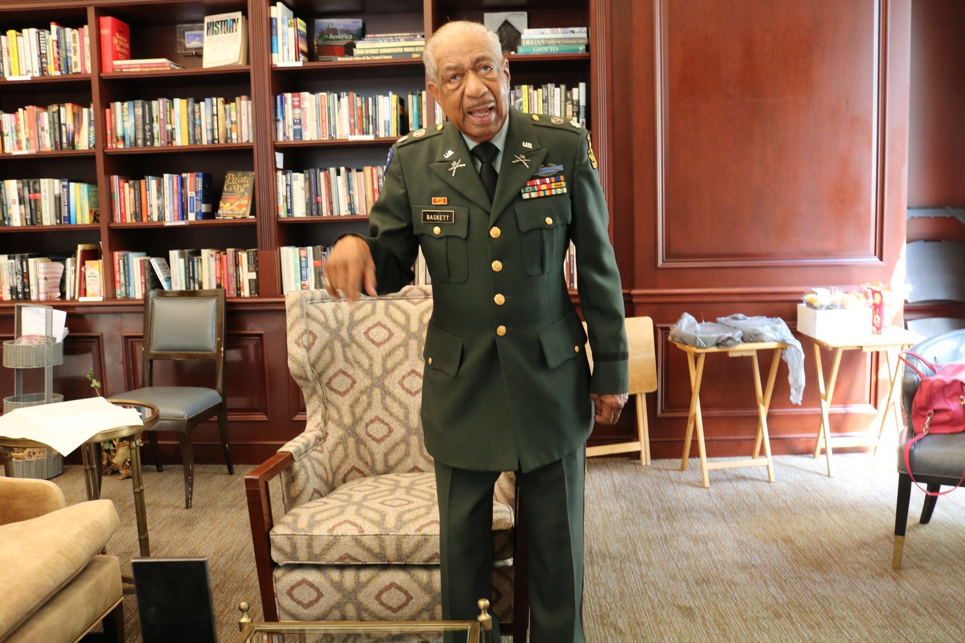Man in military uniform stands by a patterned chair in a library, pointing.