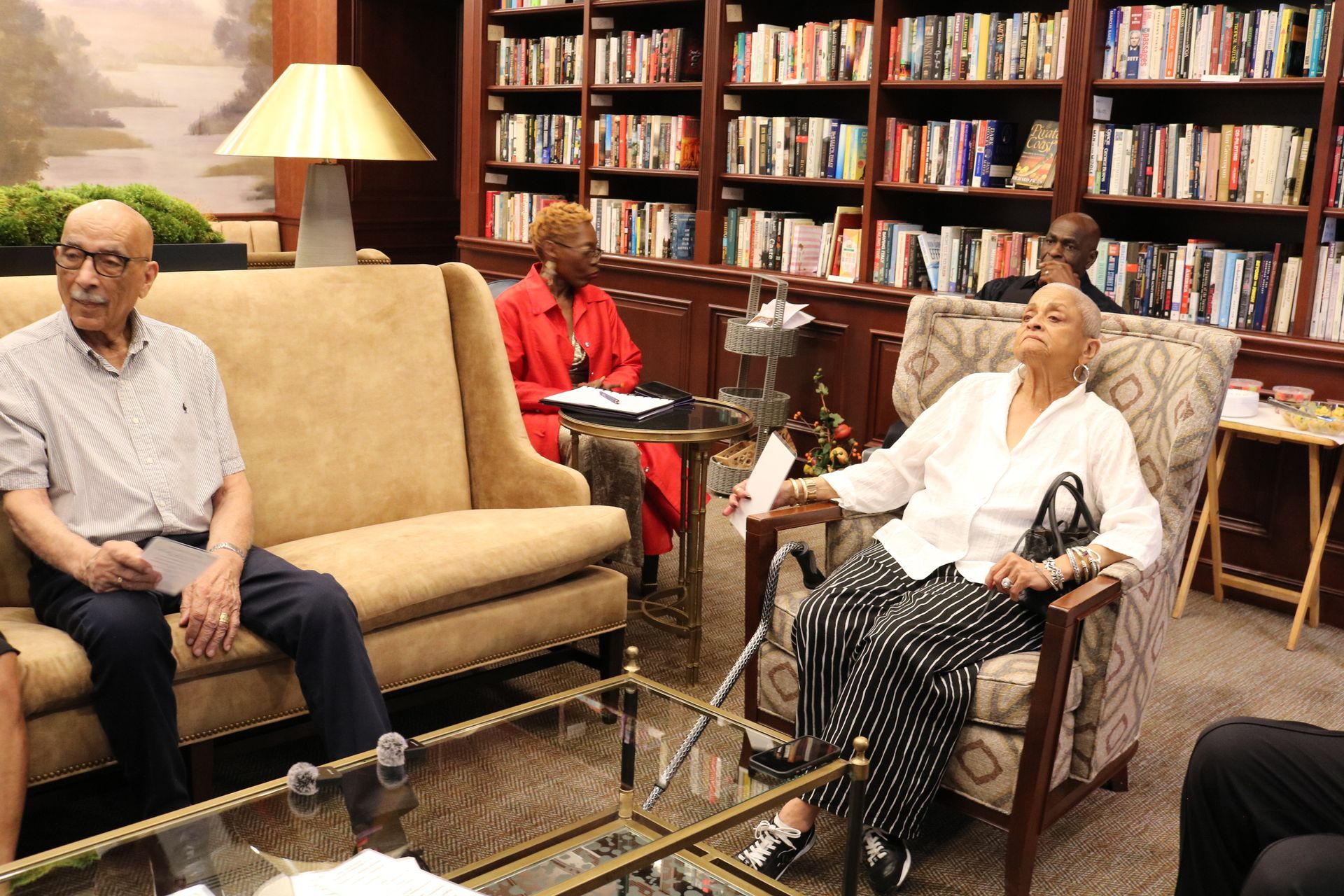 People seated in a library. One holds a cane, another a paper. Bookshelves are in the background.