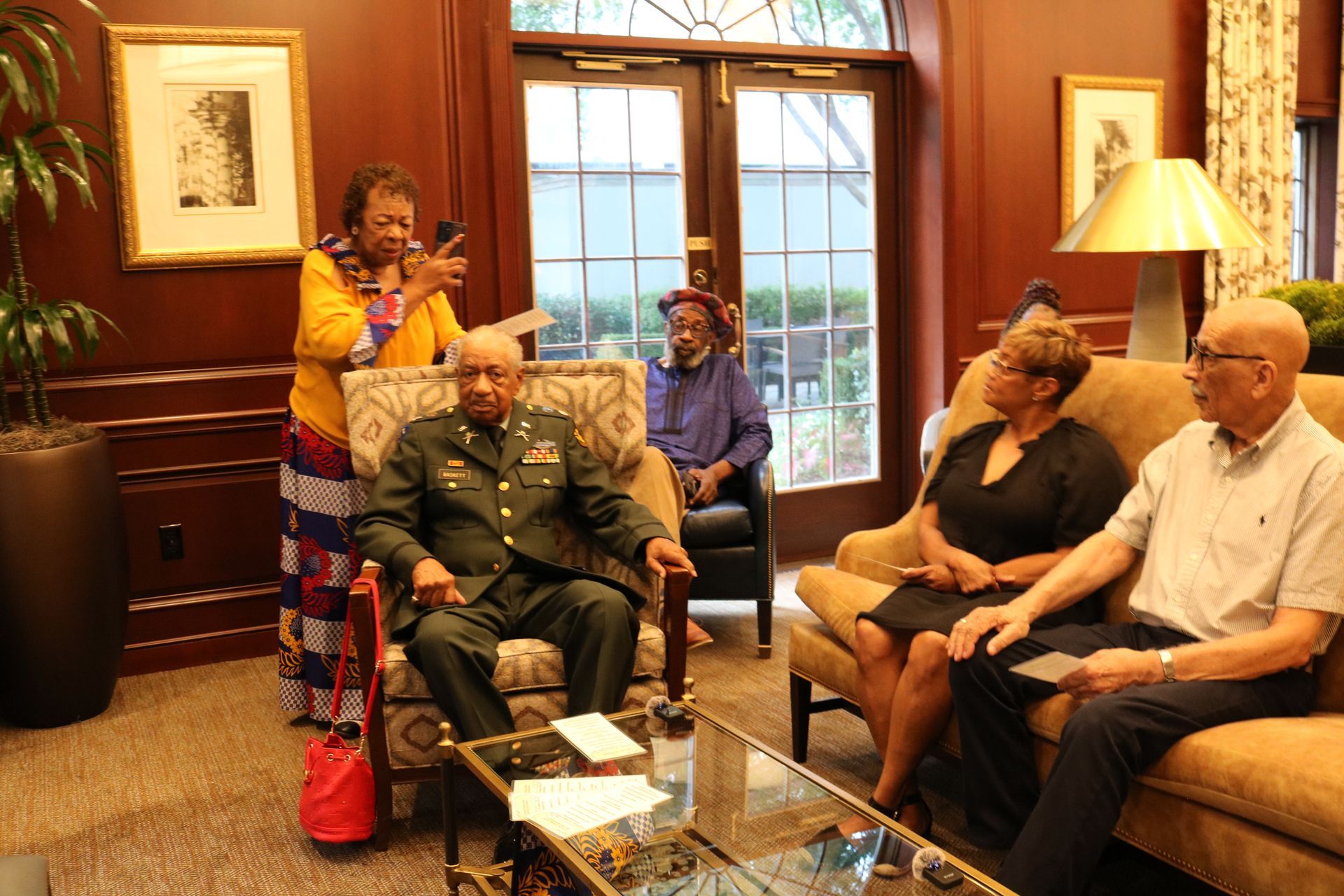 Group of people in a room; a man in military attire sits as others watch and take photos.