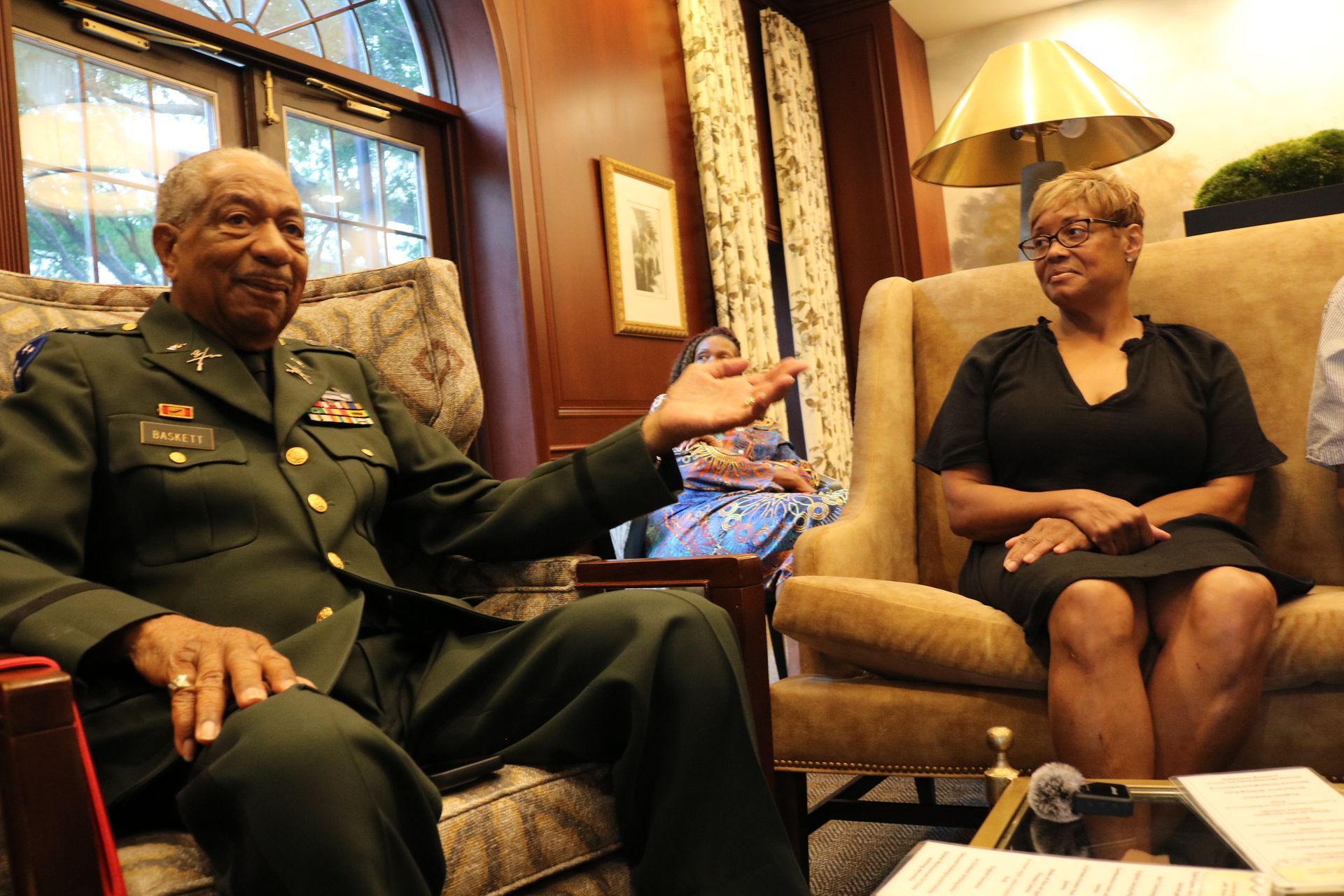 Man in military uniform gestures while conversing with a woman seated in a chair indoors.