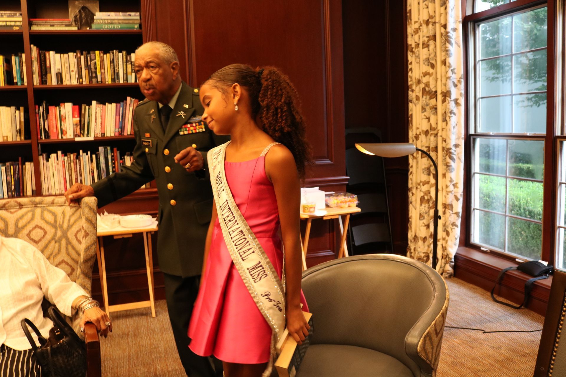 A woman in a pink dress and sash stands with a man in uniform near a bookshelf, indoors.