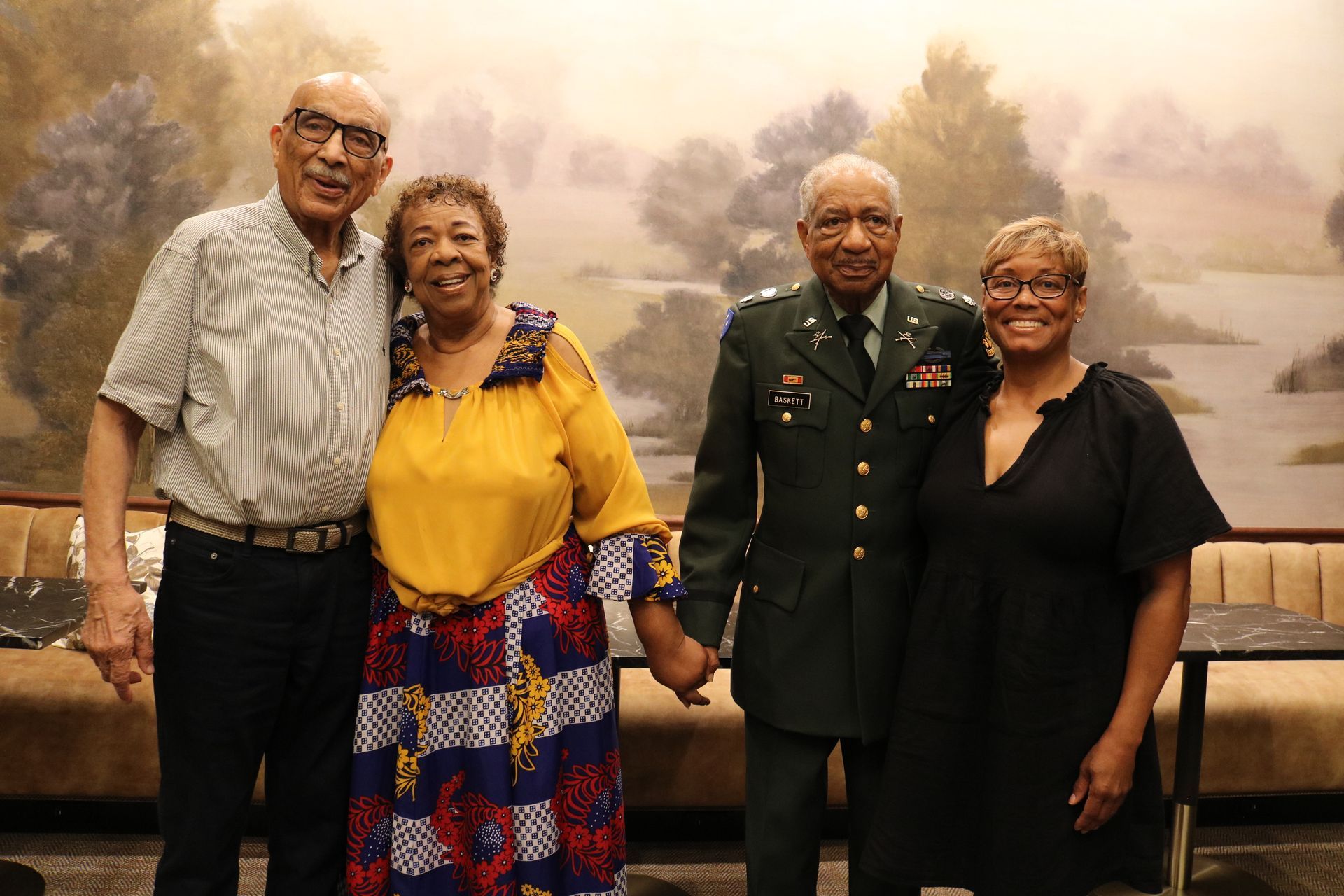 Four people smiling, holding hands, posing for photo near a mural. One in military uniform.