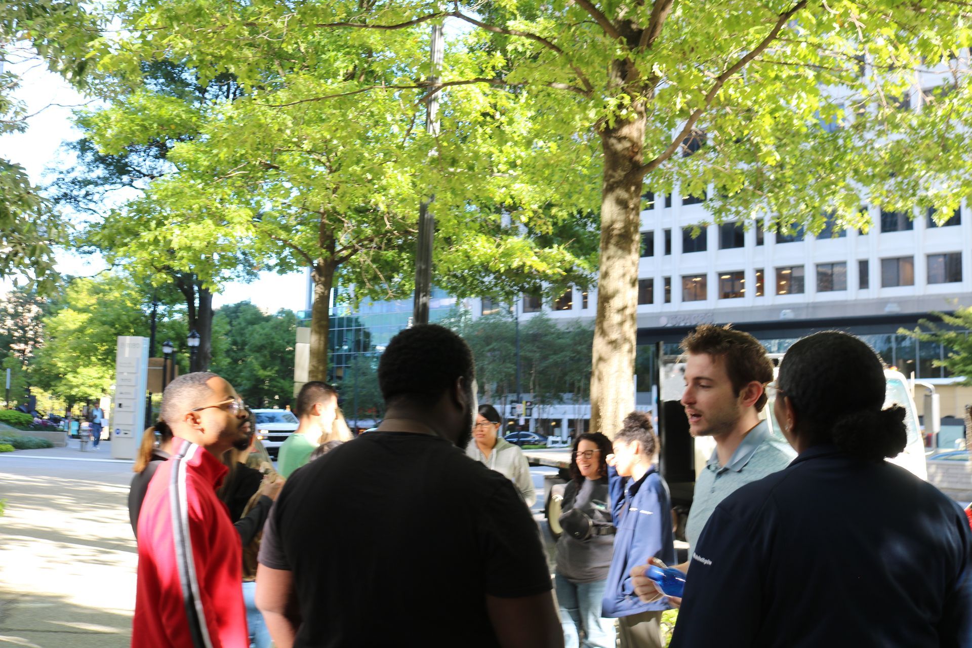 Group of people conversing outdoors near trees and a building on a sunny day.