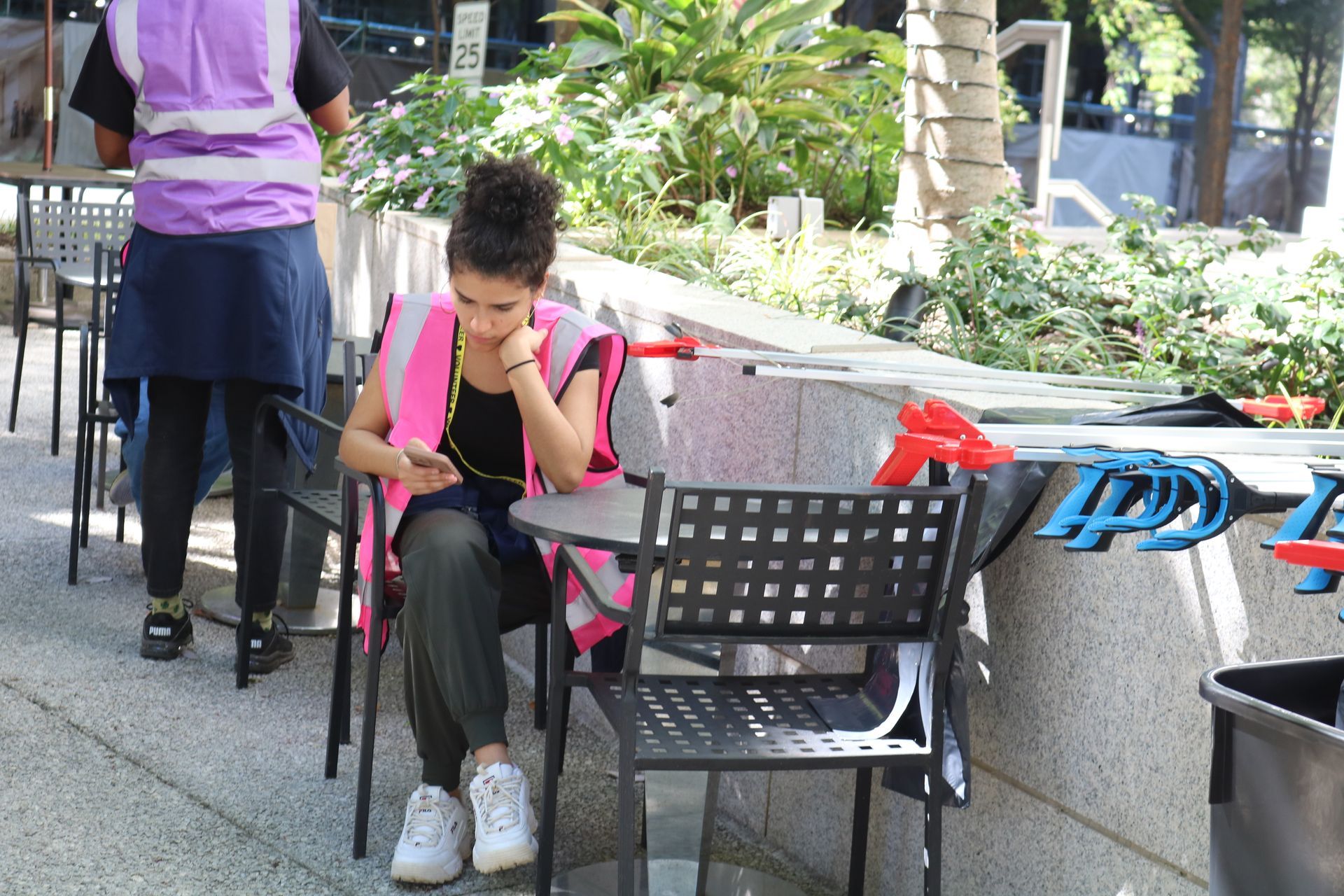Woman in a pink vest looks at her phone at a cafe table. Another person with a pink vest stands nearby.