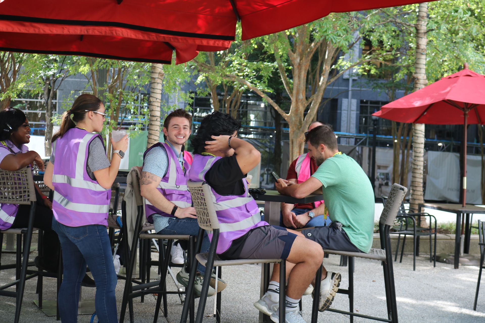 People wearing purple vests sit at outdoor tables under a red umbrella.