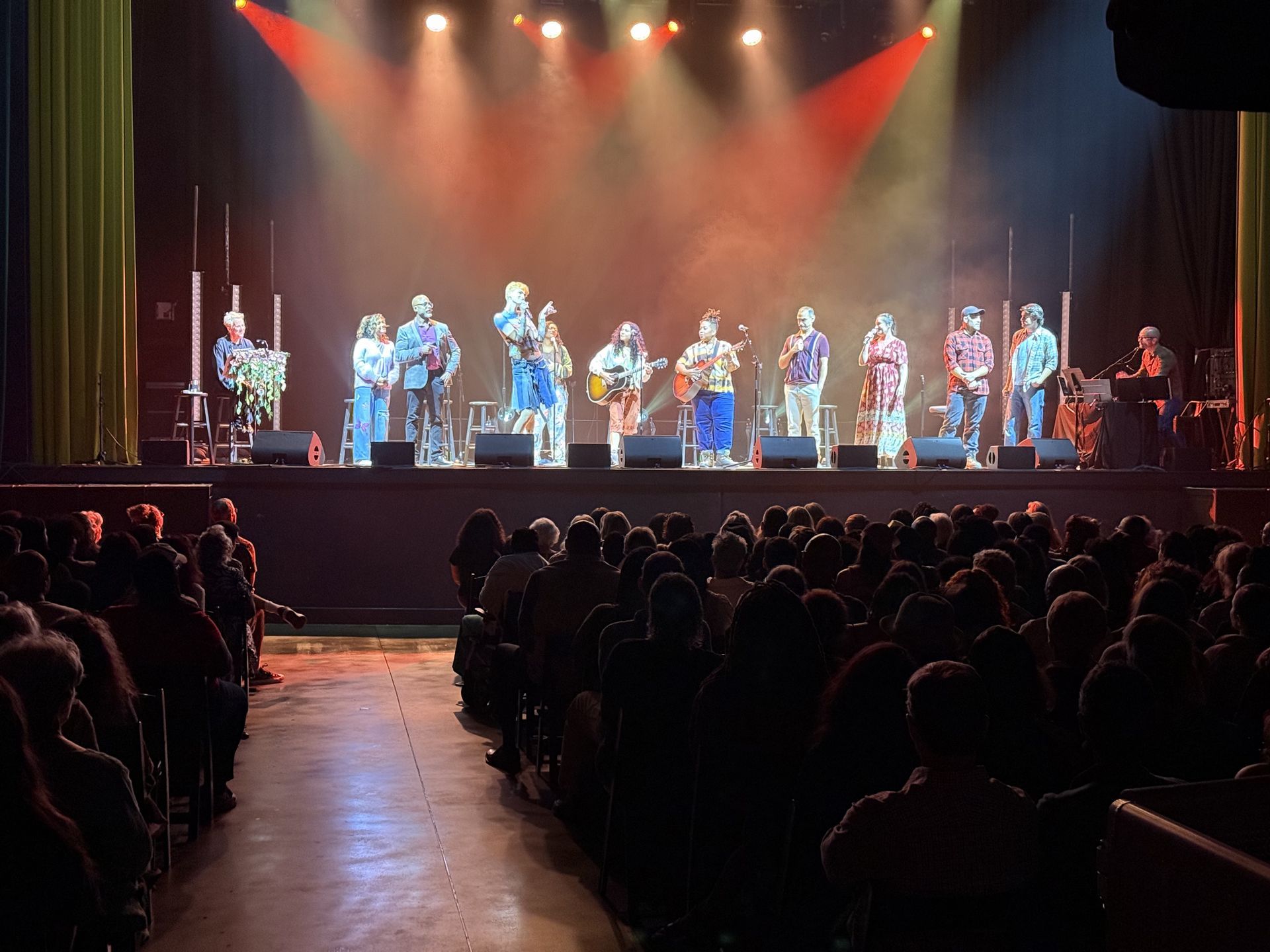 Band on stage performing for a seated audience in a theater. Stage is lit with colorful spotlights.