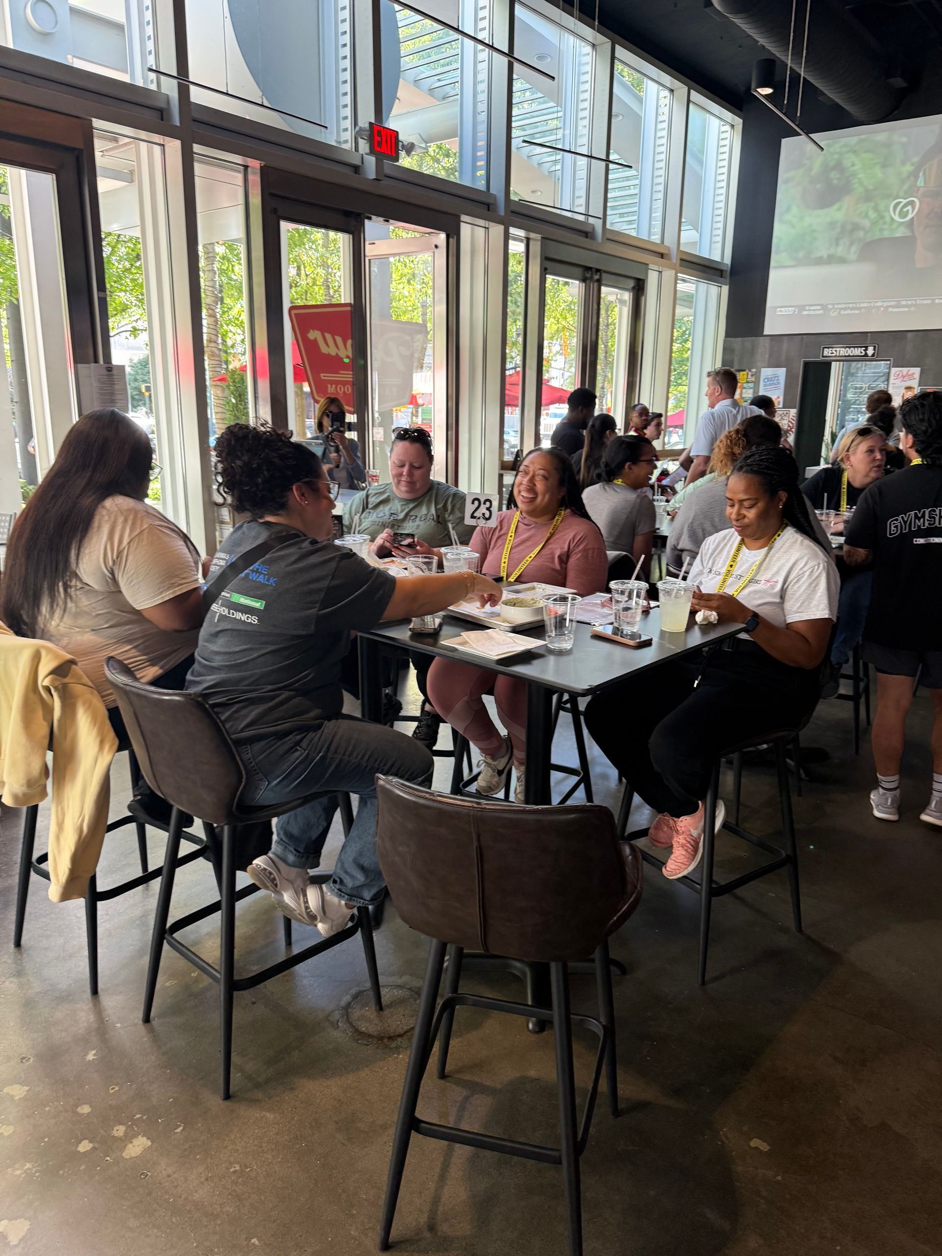 People seated around tables in a brightly lit cafe; some are working, others are talking. Large windows offer outside views.