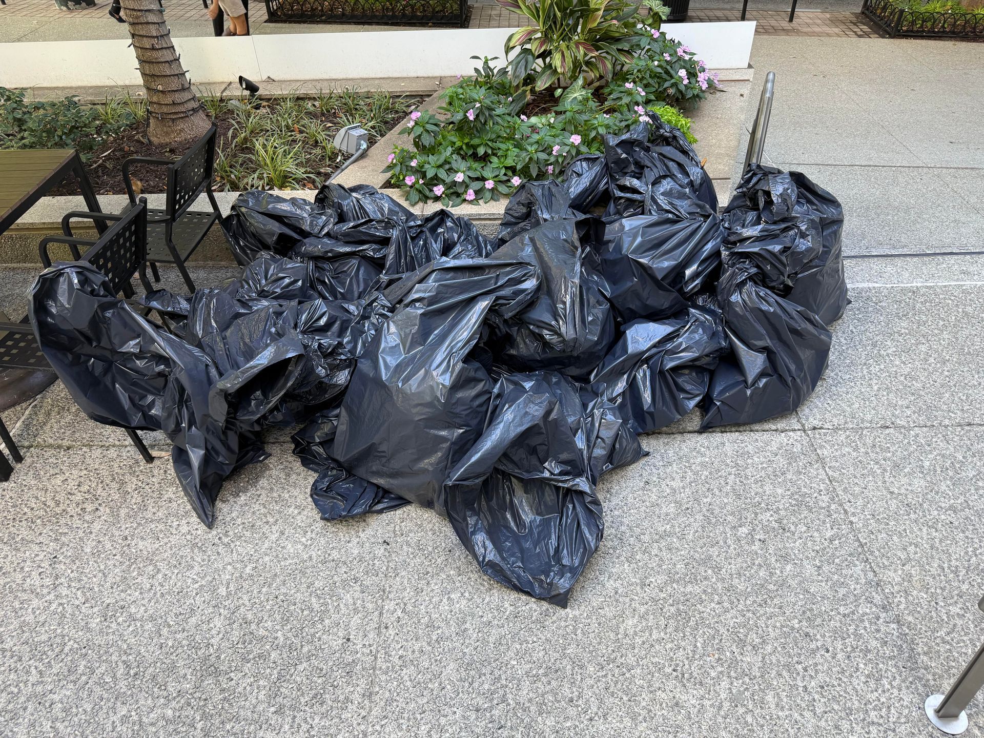 Pile of black trash bags on a stone ground near a building and some plants.