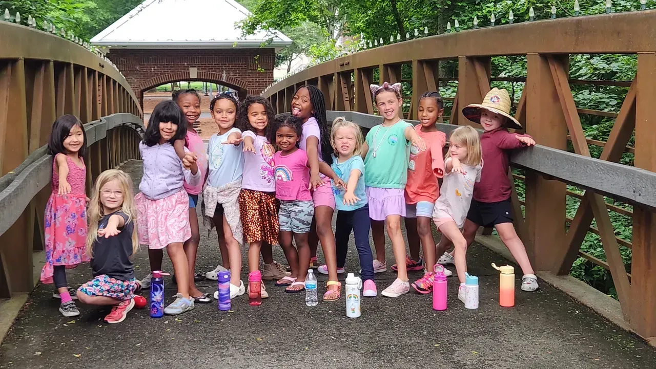 Group of children on a wooden bridge, posing with water bottles. Green trees in background.