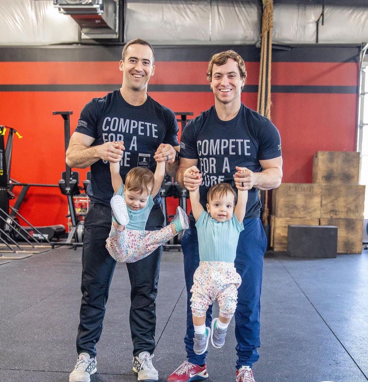 Two men are holding two children in a gym and one of them is wearing a shirt that says compete for a cure