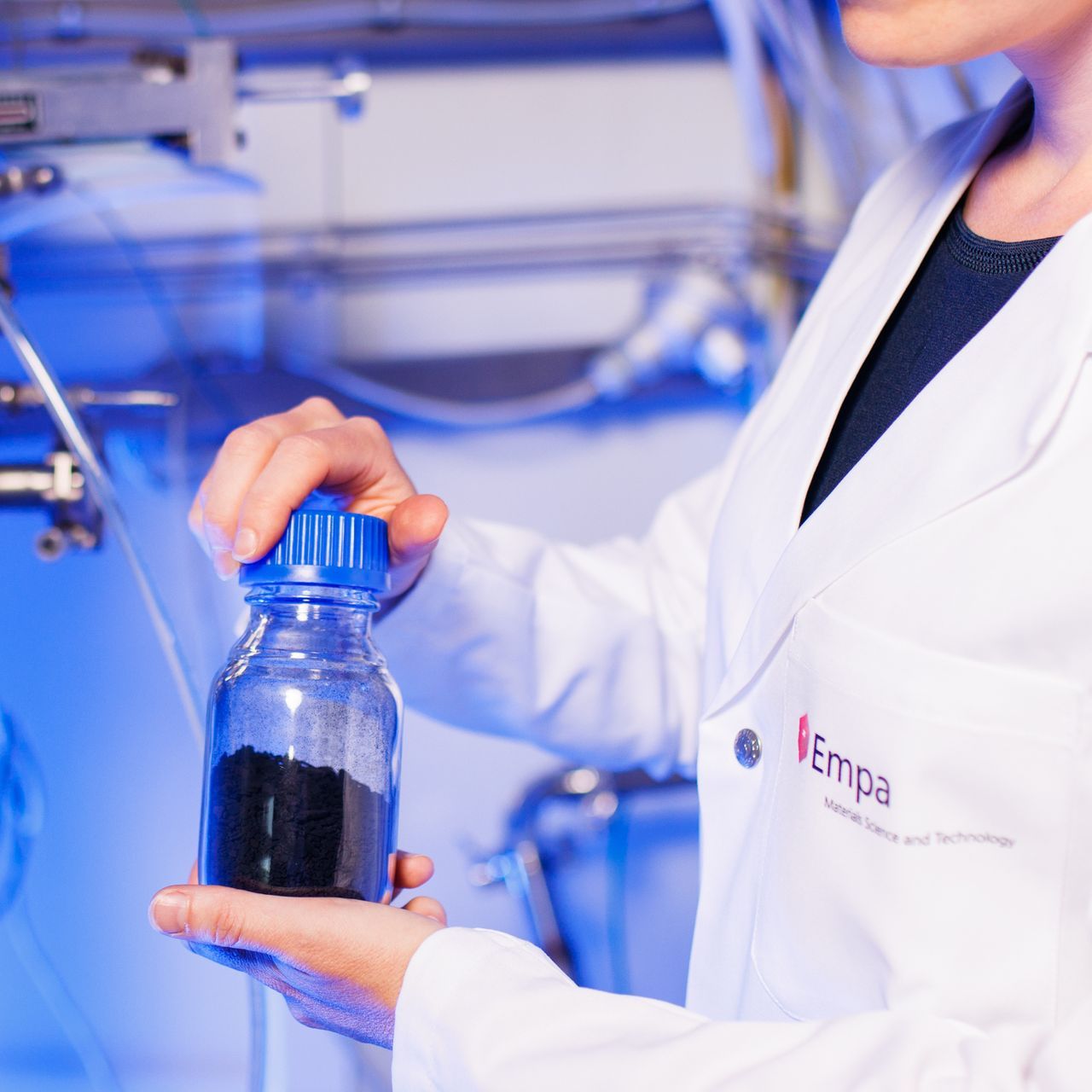 Eine Frau im Laborkittel hält eine Flasche schwarzes Pulver in der Hand - Verein zur Dekarbonisierung der Industrie