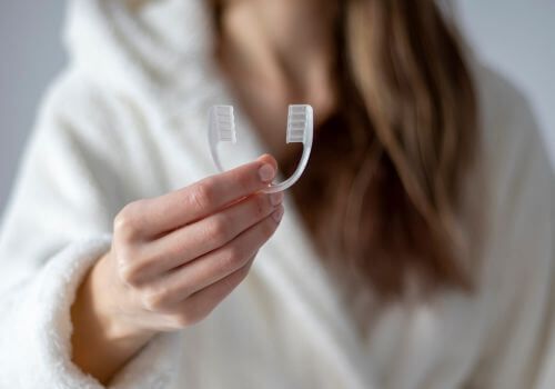 Hand holding a transparent dental aligner in front of a blurred white sweater background