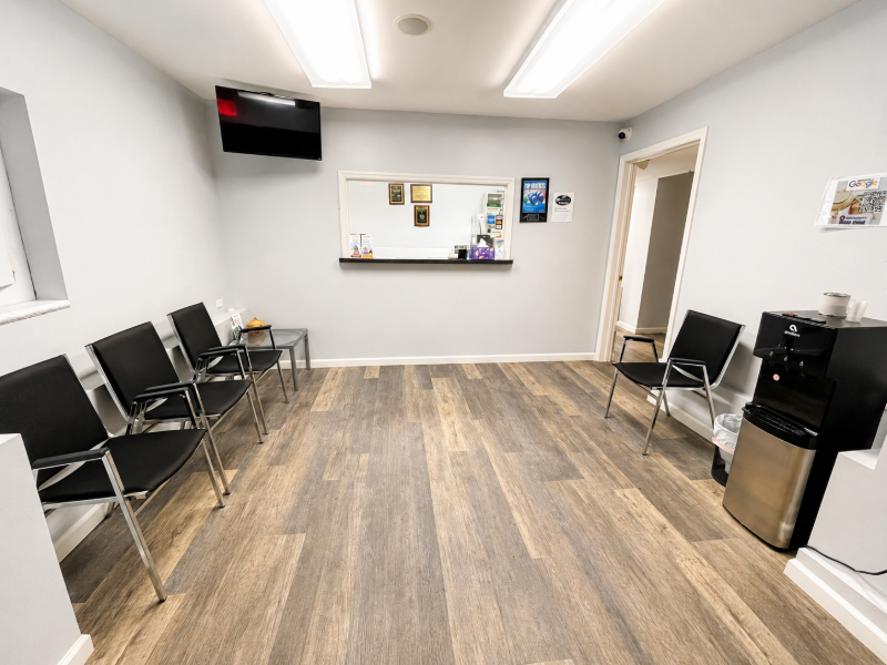 Empty waiting room with black chairs, wood-look flooring, and a reception window on the far wall