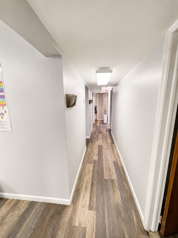 Narrow hallway with wood-look flooring, white walls, and ceiling lights leading to a doorway.