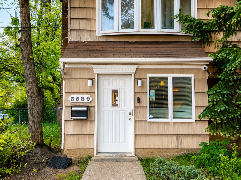 Front entrance of a tan house with white door, window, mailbox, and number 3589 amid greenery