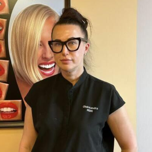 Woman in black scrubs and glasses standing beside a wall portrait with a large smiling face.