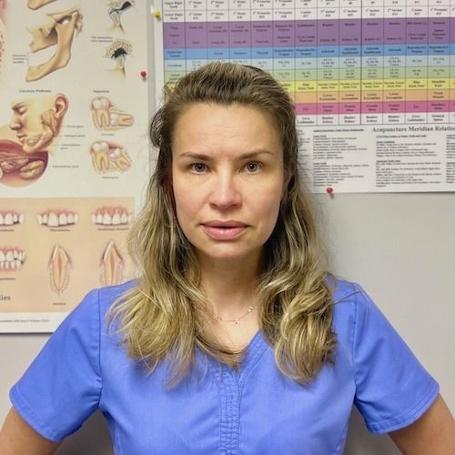 Woman in blue scrubs standing in a dental office, with a smile chart and anatomical posters behind her.
