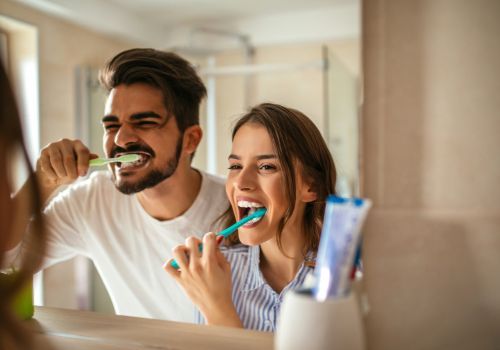 Two people brushing their teeth together in a bathroom, smiling at a mirror