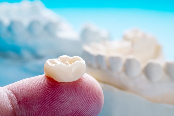 A tooth-shaped dental crown balanced on a fingertip, with blurred teeth and blue background behind it