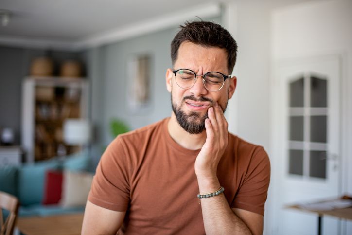 Man in a brown shirt holding his cheek, looking pained in a bright living room