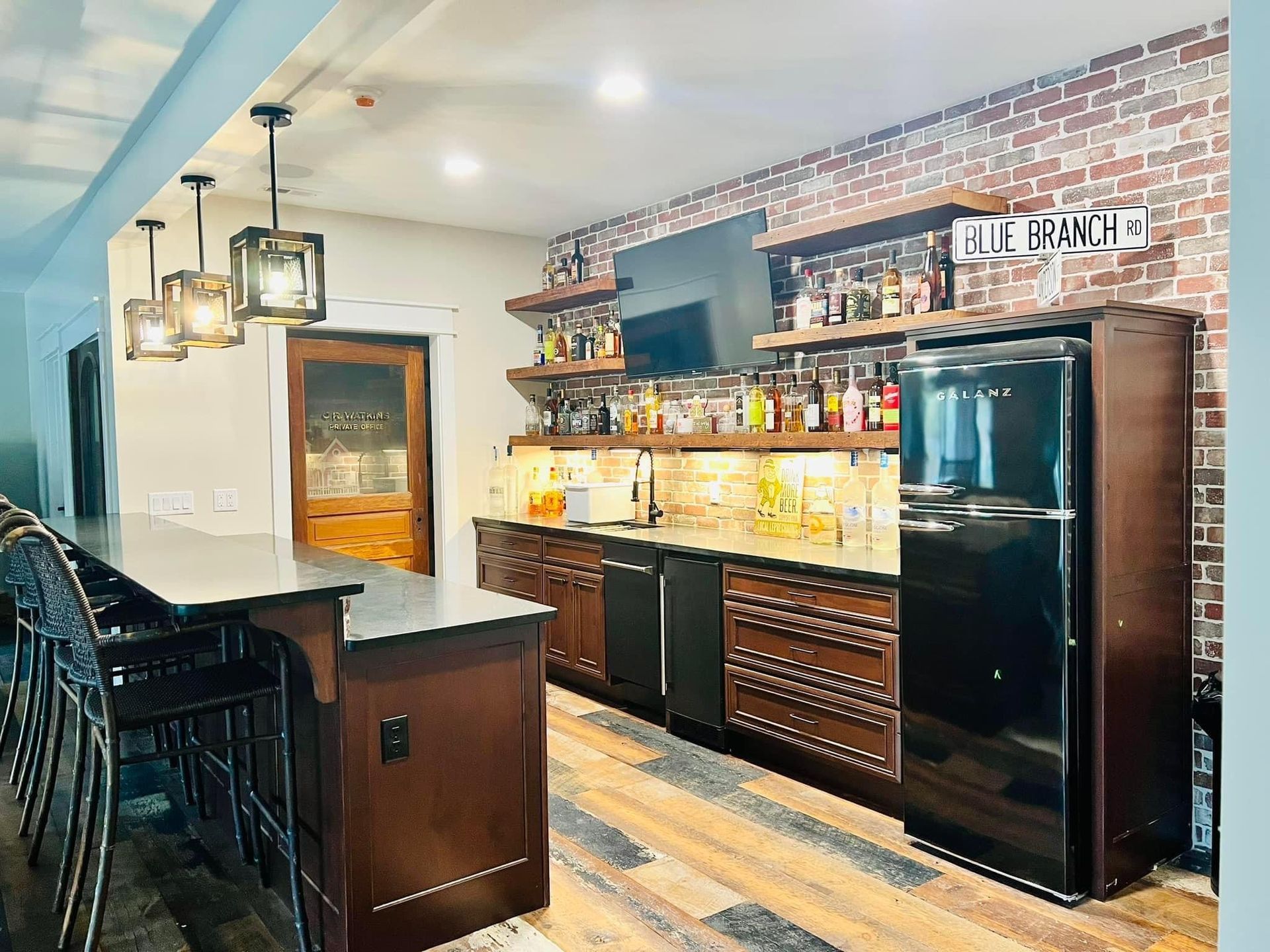 A kitchen with a large island , stools , a refrigerator , and a brick wall.