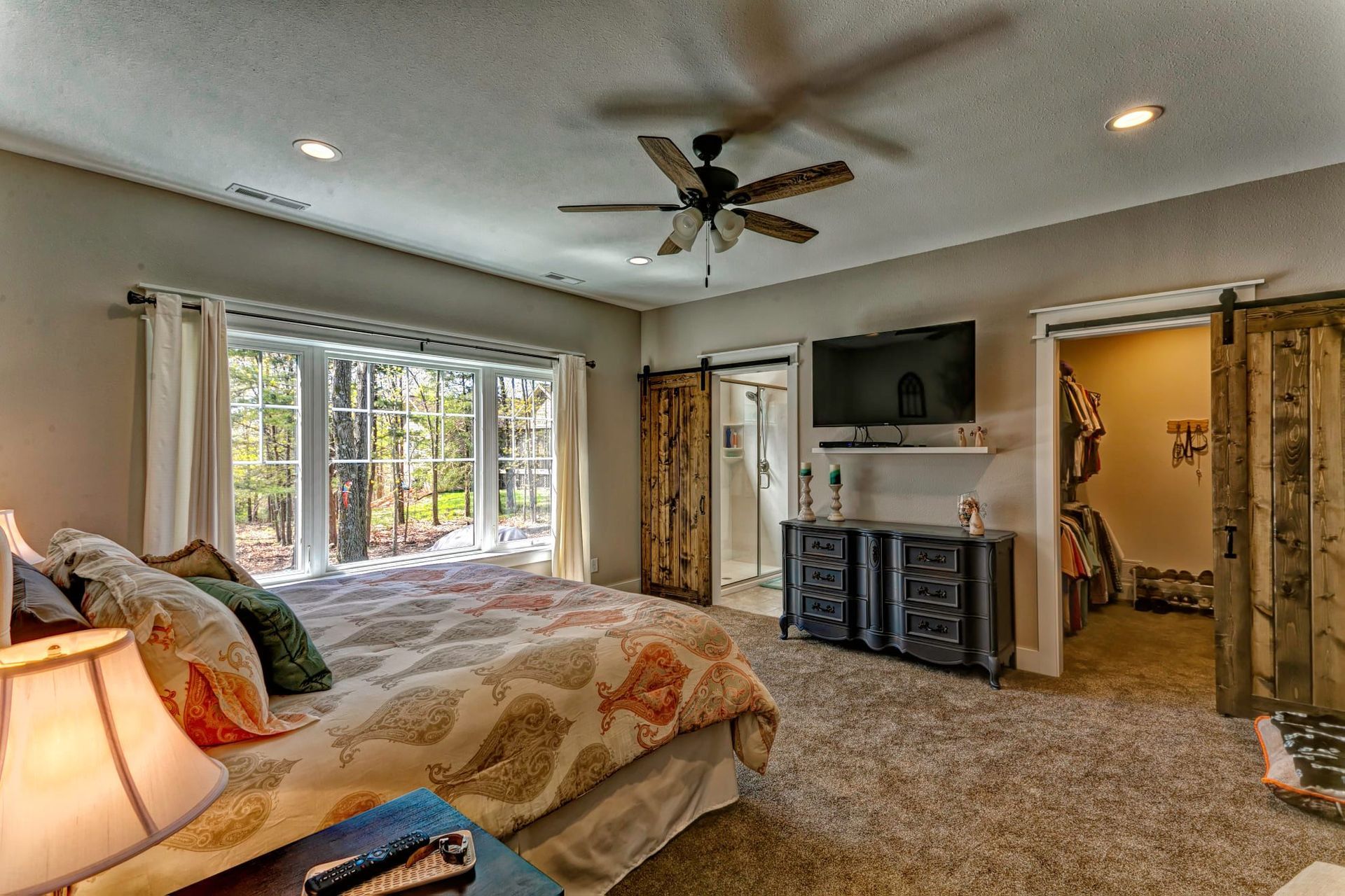 A large bedroom with a king size bed , sliding barn doors , a flat screen tv and a ceiling fan.