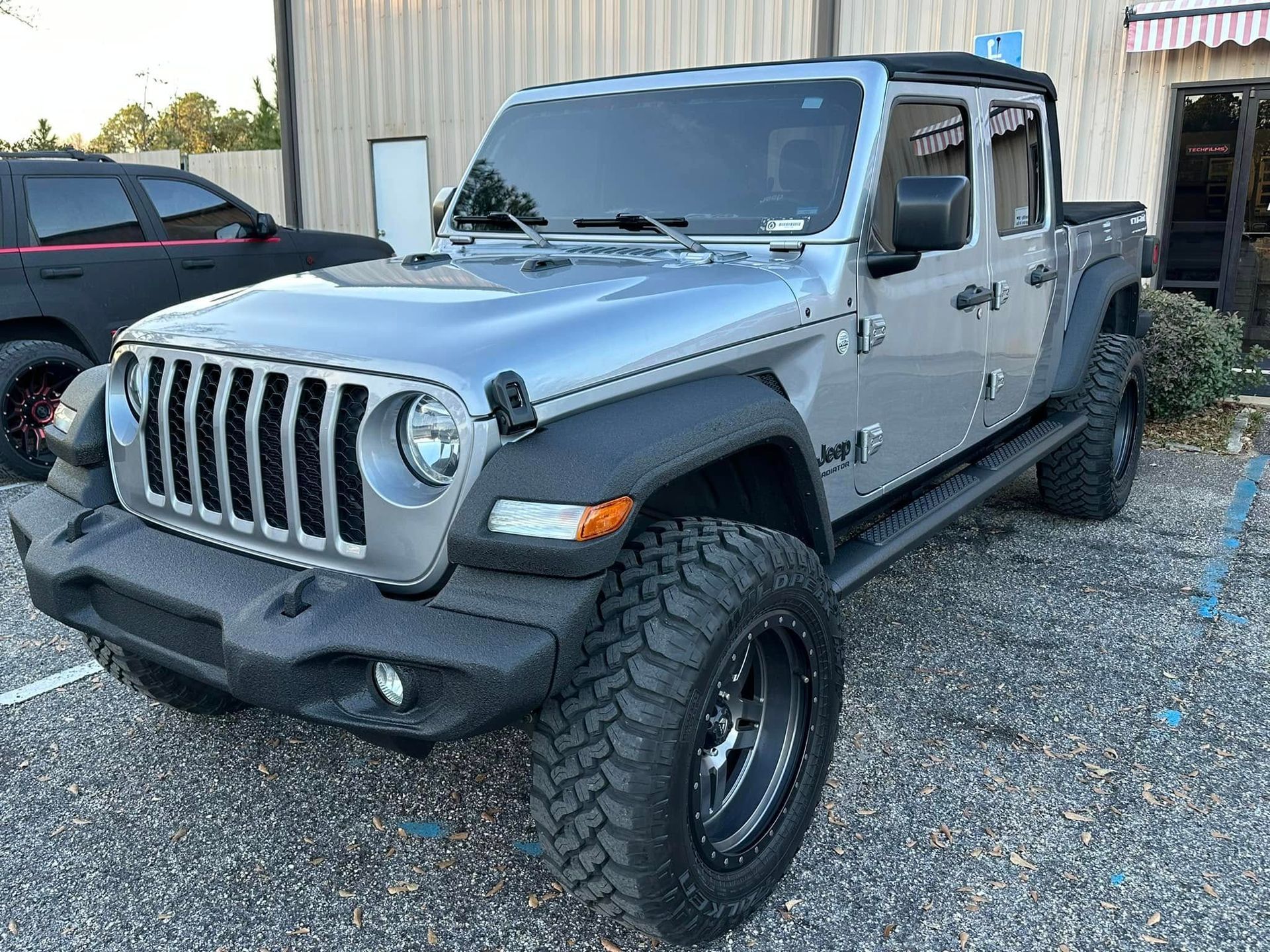 A silver jeep is parked in a gravel lot in front of a building.