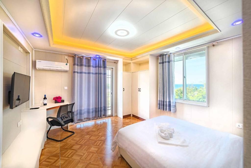 Bedroom with bed, desk, TV, and window overlooking the sea, warm lighting.