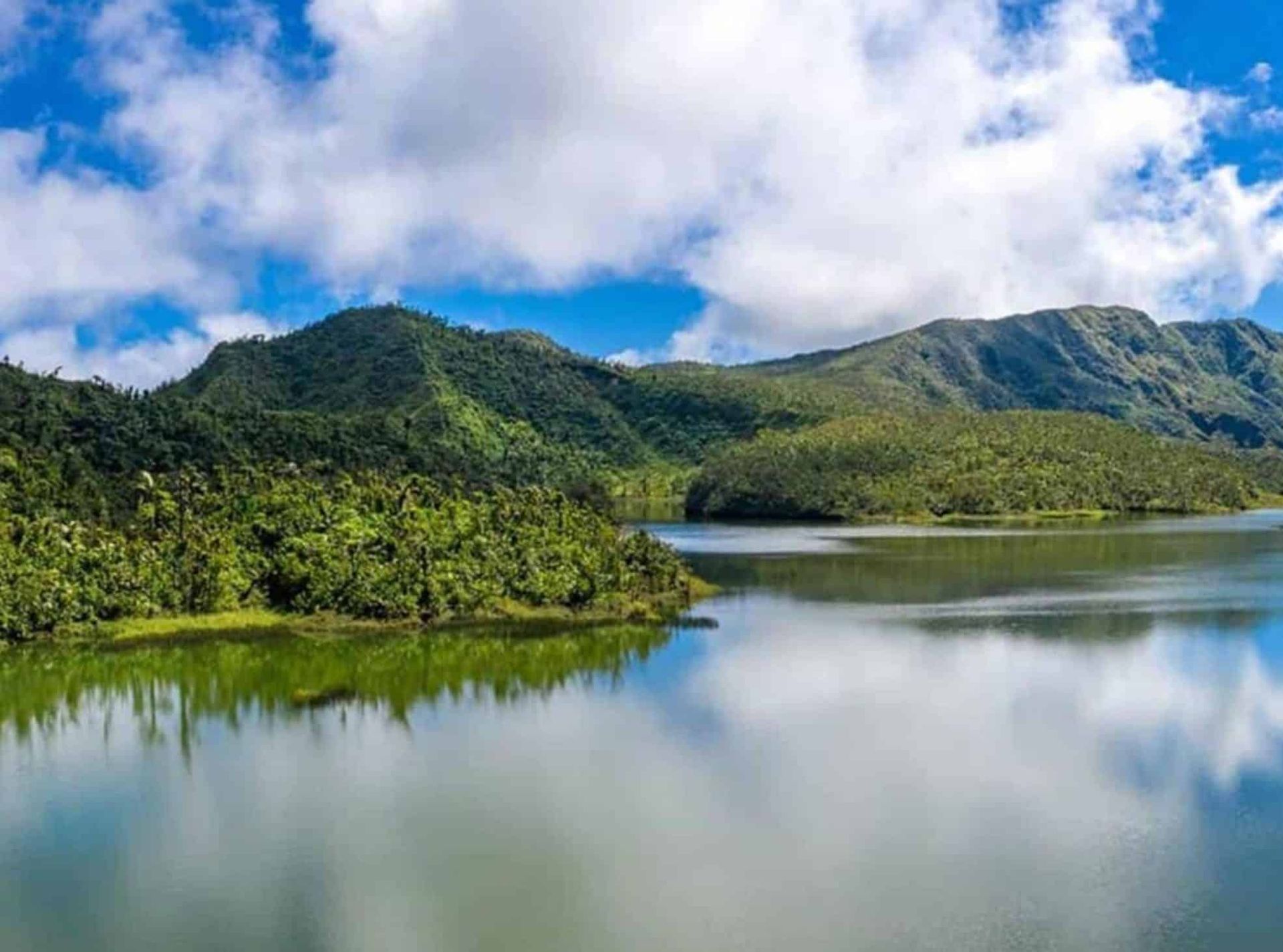 Lush green mountains reflected in a calm lake under a cloudy blue sky.